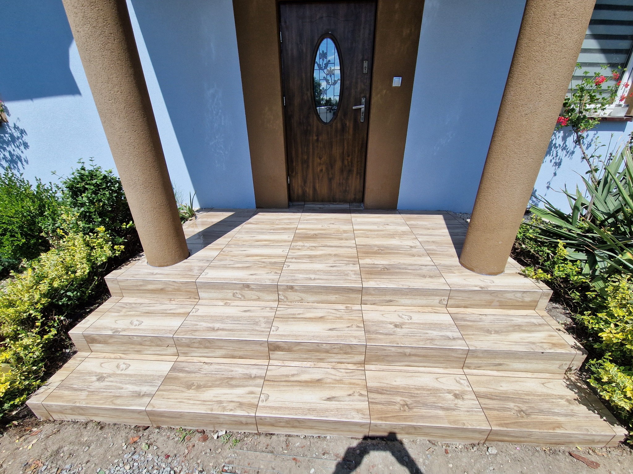 Wykończone płytkami schody wejściowe do domu, z brązowymi kolumnami po bokach i ozdobnymi roślinami. Płytki imitują drewno, dodając naturalnego charakteru.