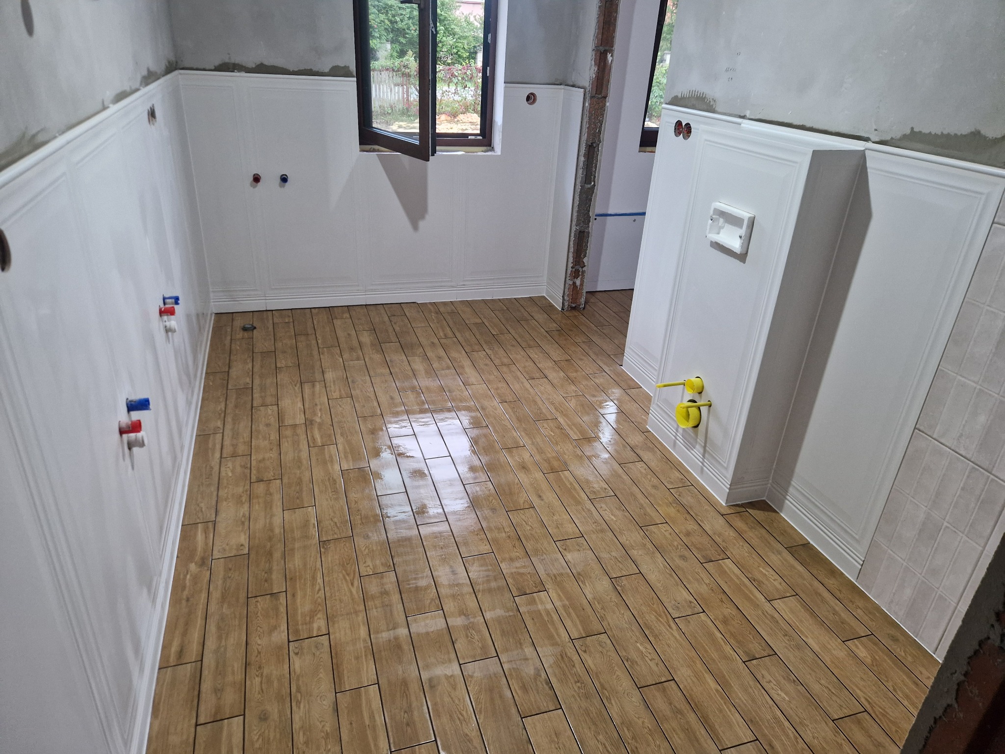 Wykończona łazienka z drewnopodobną podłogą i białymi panelami ściennymi. Widoczne przyłącza wodne i okno z widokiem na zieleń. Częściowo wykończone ściany.