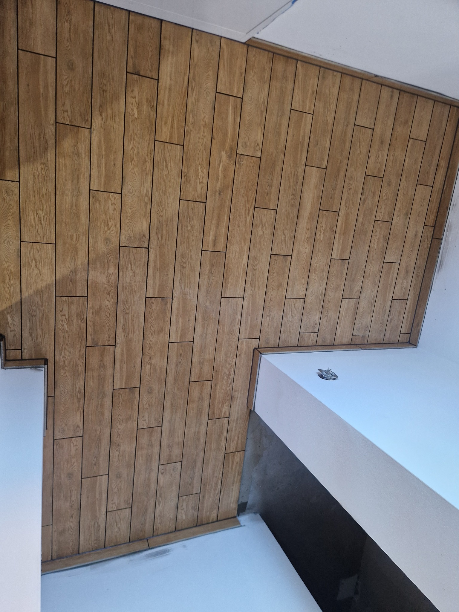 Ukończona ściana z paneli imitujących drewno, ułożonych w pionowy wzór. Widoczne fragmenty białych ścian i sufitu. Nietypowy kąt ujęcia.