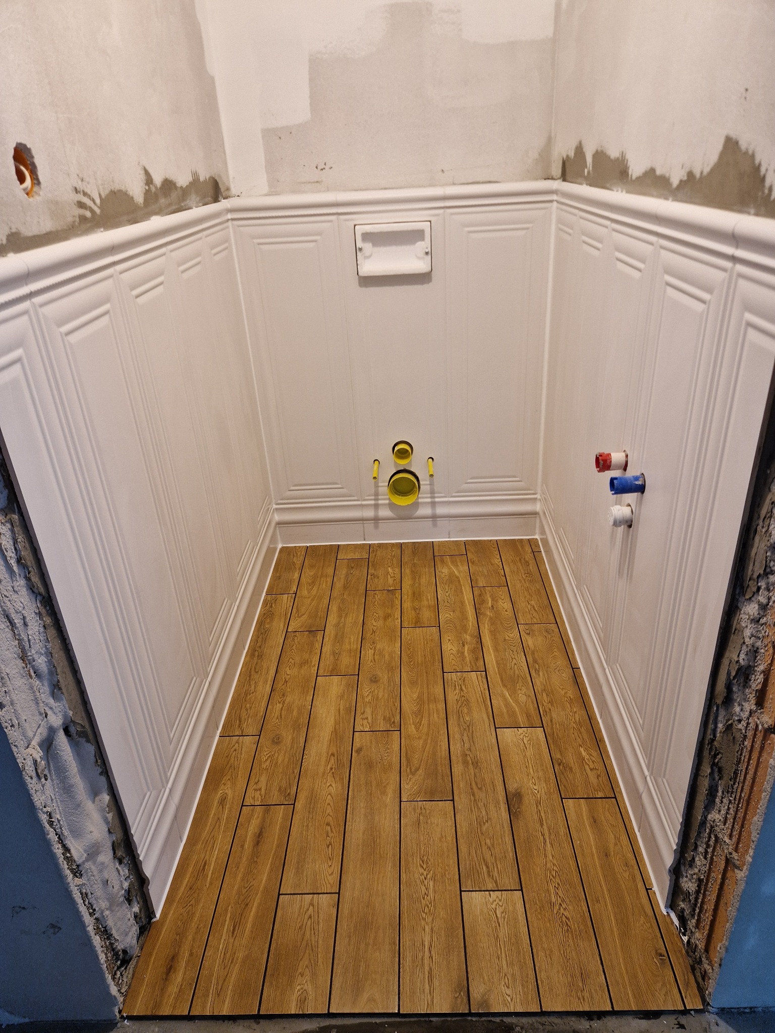 Wnętrze łazienki w trakcie prac: ułożona podłoga z płytek imitujących drewno, białe panele ścienne z dekoracyjnymi listwami, widoczne instalacje wod-kan.