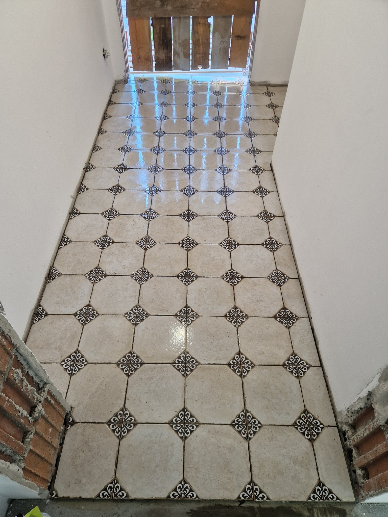 Wykończona podłoga z płytek w kremowym odcieniu z dekoracyjnym wzorem, widok z góry na fragment pomieszczenia z białymi ścianami i drewnianym progiem.