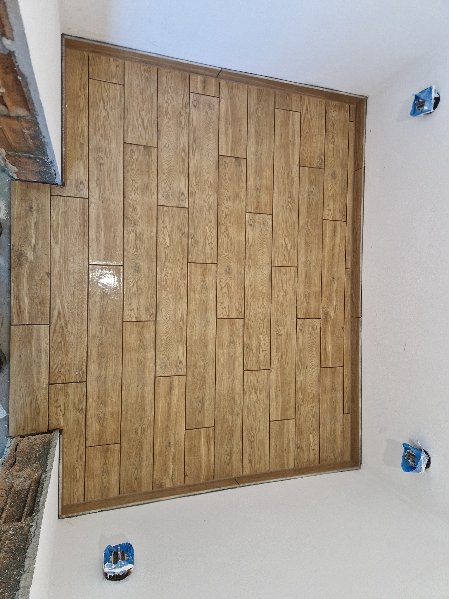 Podłoga wyłożona brązowymi płytkami imitującymi drewno. Widoczne fragmenty ścian i wystające puszki elektryczne. Ułożenie w jodełkę, z połyskiem na niektórych płytkach.