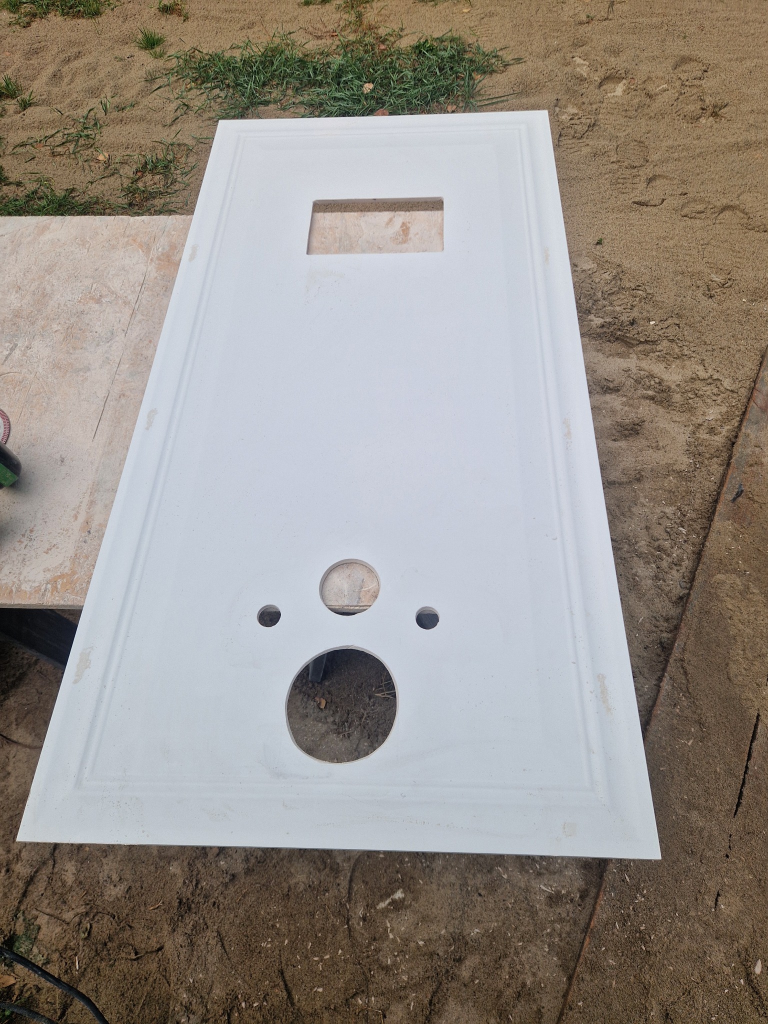 Biały panel z wyciętymi otworami leży na zewnątrz, prawdopodobnie przygotowany do montażu w łazience. Widoczne otwory na umywalkę i armaturę.