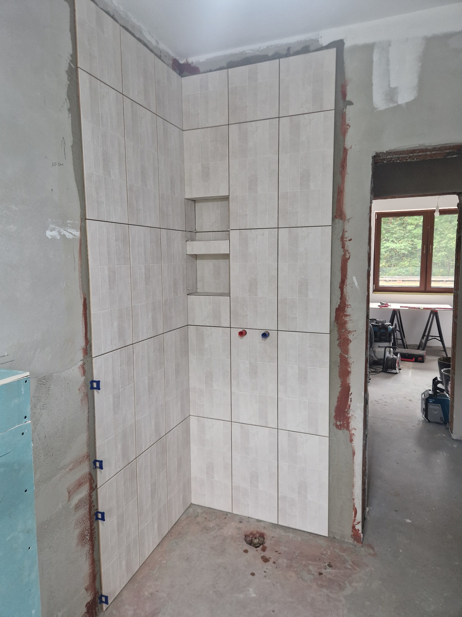 Wnętrze łazienki w trakcie remontu. Ułożone beżowe płytki na ścianie z wnękami. Widoczne nieotynkowane ściany, narzędzia i okno w tle. Częściowo ukończona praca glazurnicza.