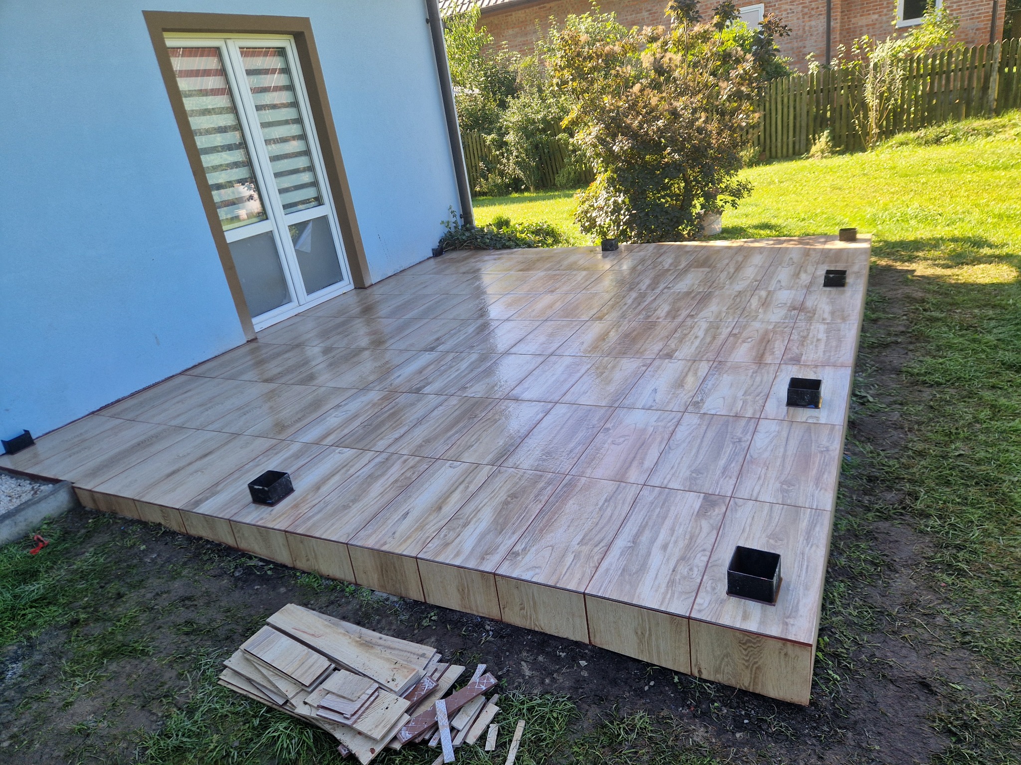 Wykończony taras z płytek imitujących drewno w Nowosielcu, z widocznymi elementami konstrukcyjnymi i resztkami materiałów budowlanych na trawniku.