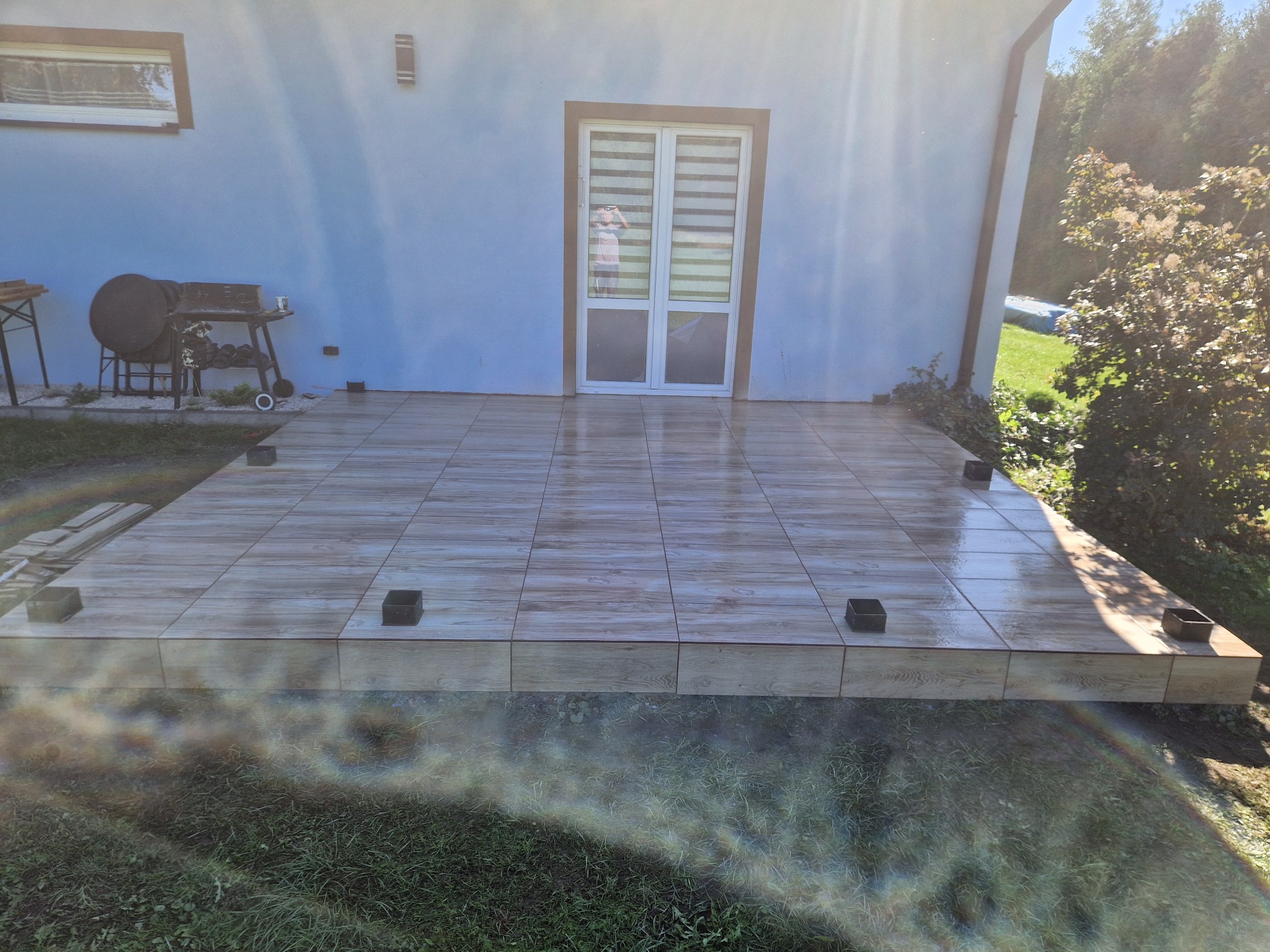 Wykończony taras przed domem, wyłożony płytkami imitującymi drewno. Widoczne czarne podpory pod konstrukcją. W tle ściana budynku i zieleń ogrodu.