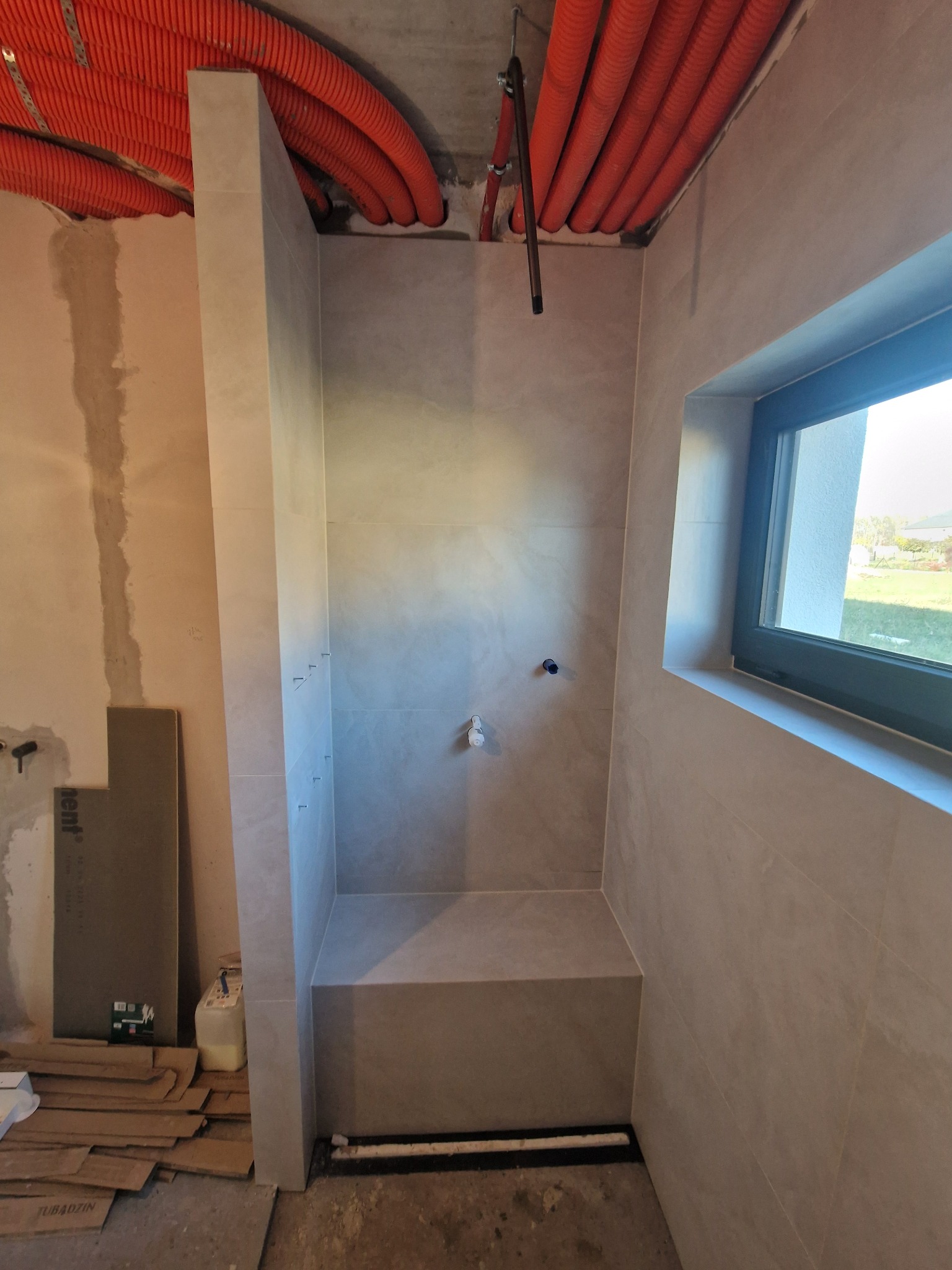 Wykończona kabina prysznicowa z jasnymi płytkami, widoczne rury na suficie i okno z widokiem na zieleń. Minimalistyczny design, surowy stan wykończenia poza kabiną.