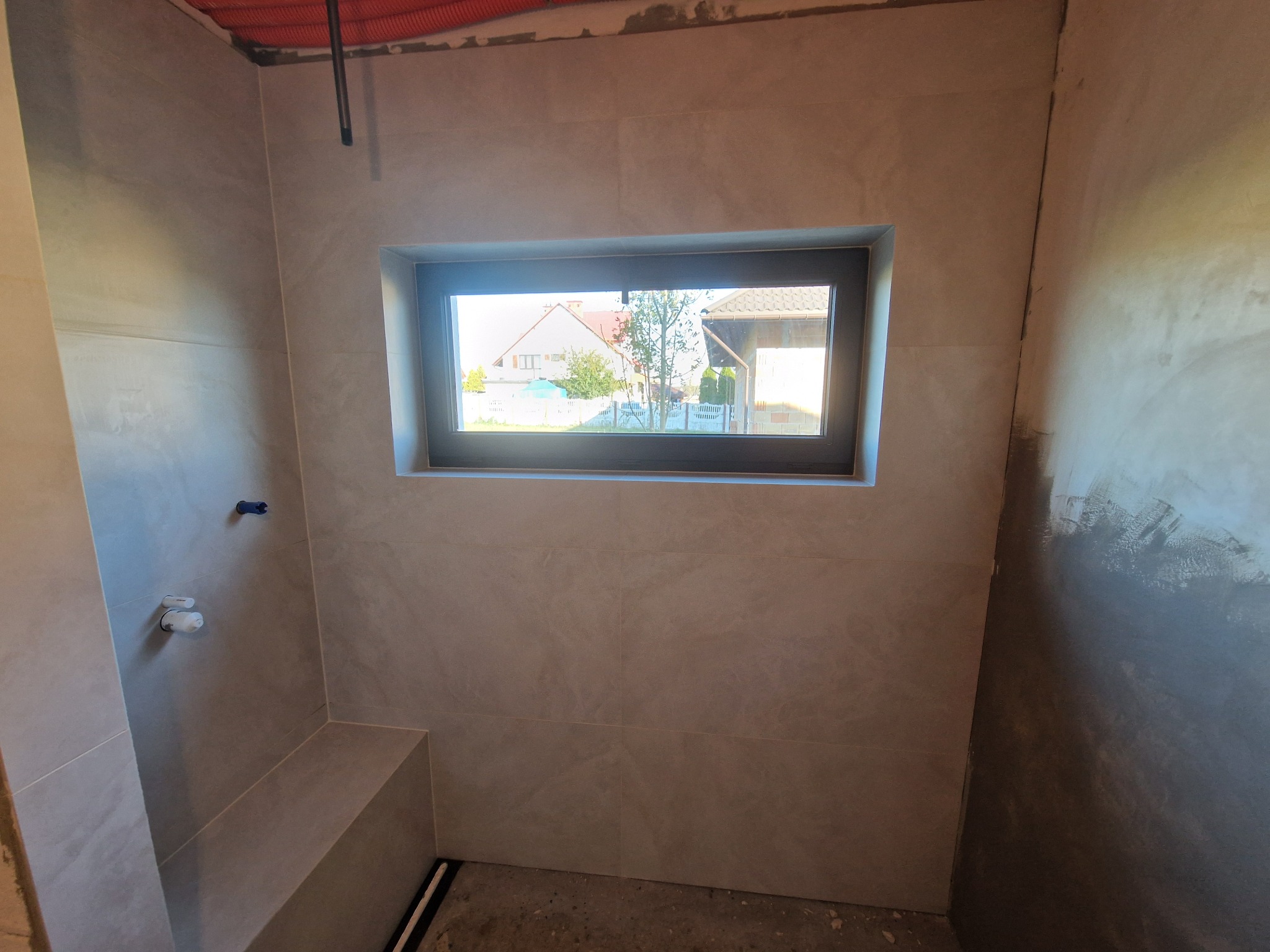 Wnętrze łazienki w trakcie wykańczania: szare płytki na ścianach, okno z widokiem na domy, instalacja sanitarna częściowo widoczna. Surowy stan podłogi.