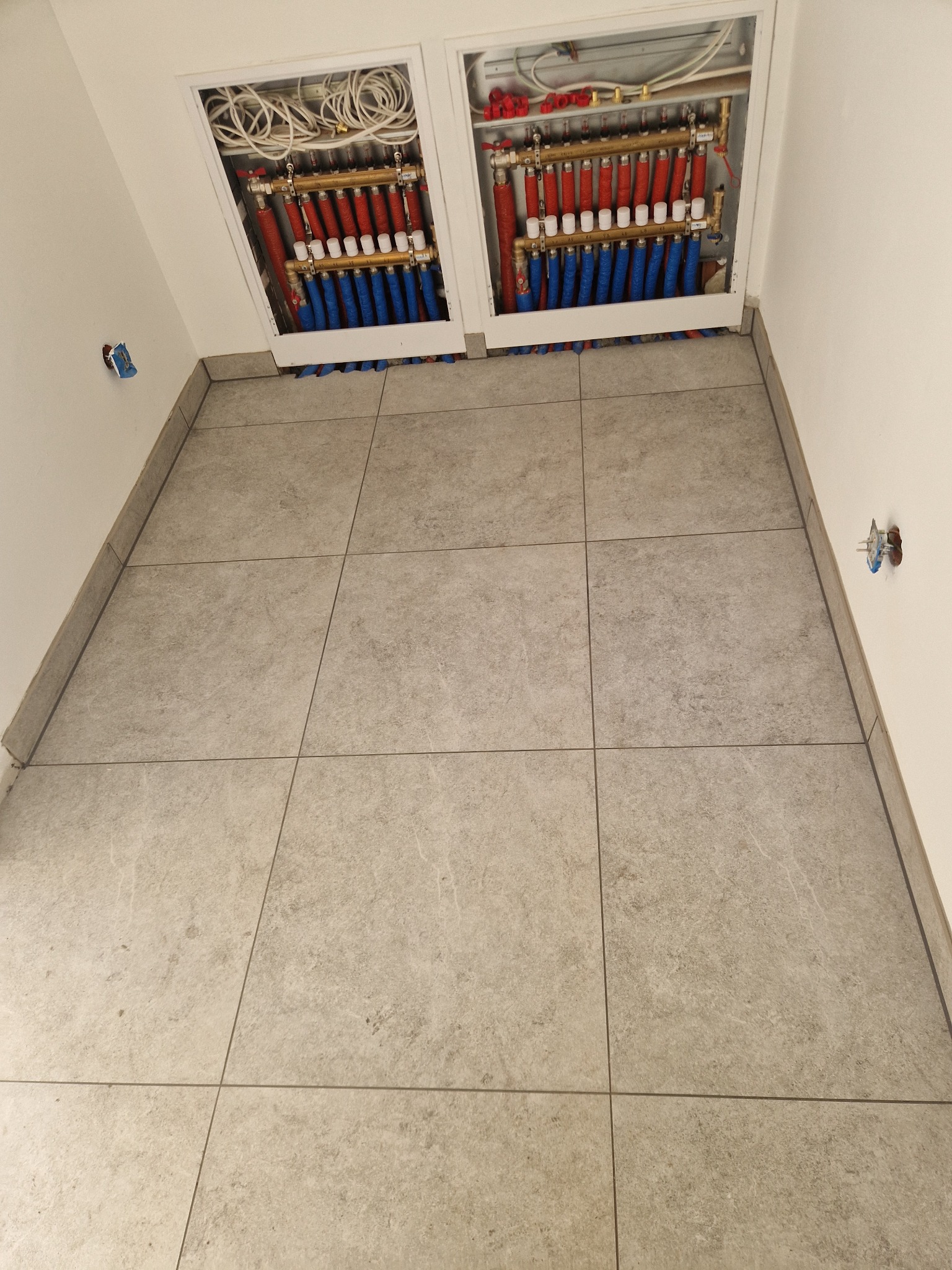 Szare płytki podłogowe w łazience w Nowosielcu. Widoczne dwie szafki z instalacją ogrzewania podłogowego z rurami w kolorze czerwonym i niebieskim.