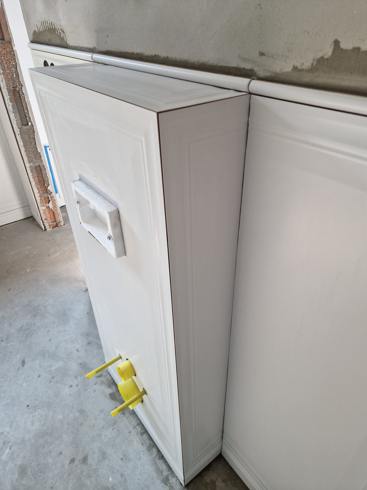 Biała obudowa stelaża WC z wyprowadzonymi żółtymi rurkami instalacji wodnej. Widoczna puszka podtynkowego gniazda elektrycznego i fragment ściany z surowym tynkiem.