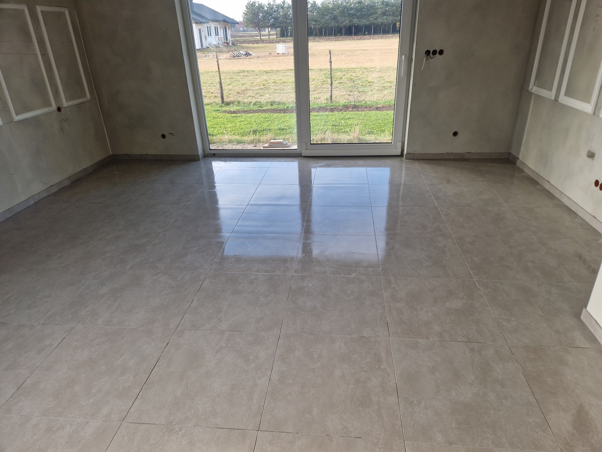 Jasne, błyszczące płytki podłogowe w nowym wnętrzu z dużym oknem. Ściany w surowym wykończeniu, widoczne puszki elektryczne. Na zewnątrz widok na pole i zabudowania.