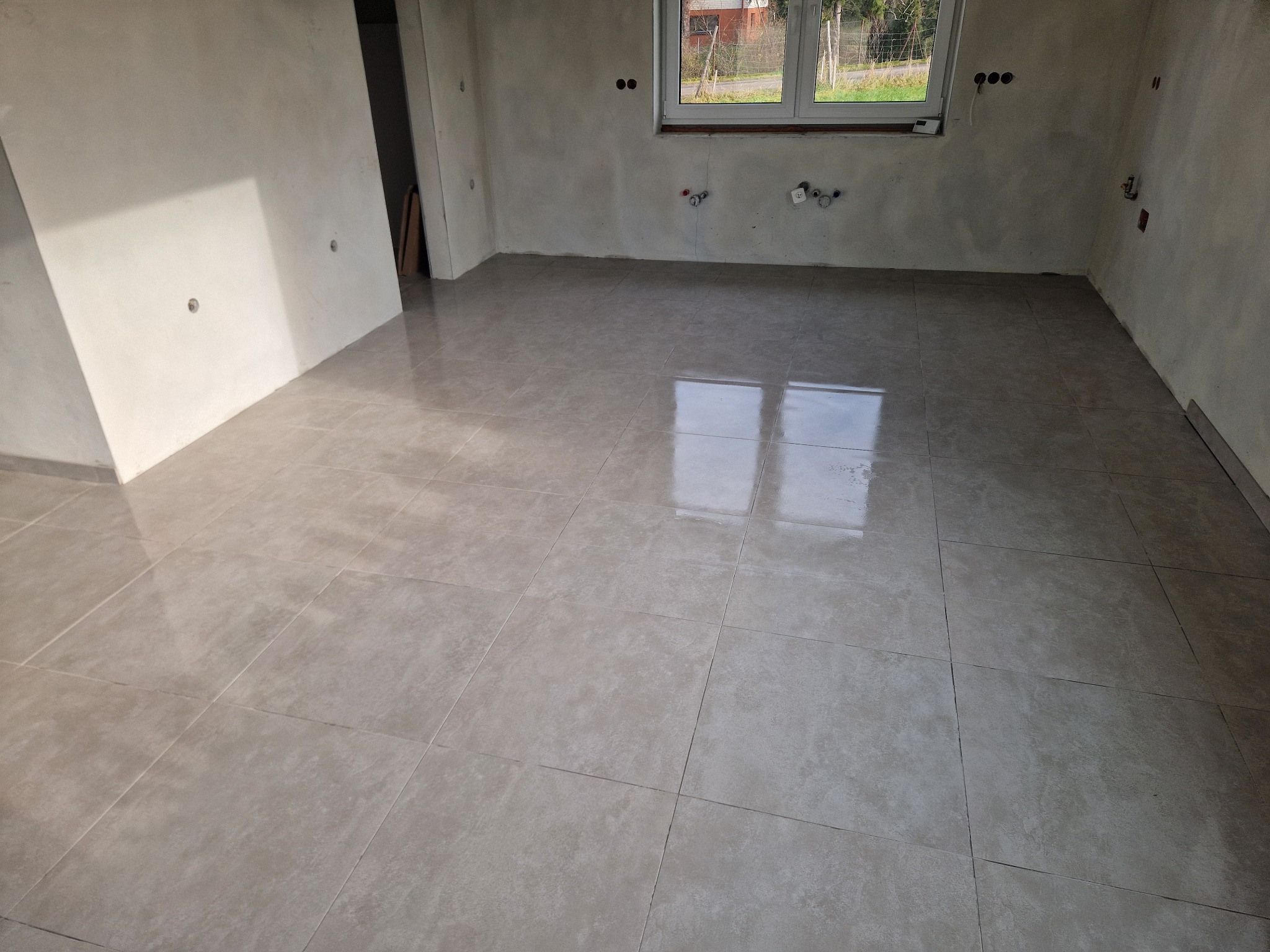 Jasne, kwadratowe płytki podłogowe z widocznymi fugami w surowym wnętrzu z oknem. Ściany w stanie deweloperskim, widoczne instalacje. Minimalistyczny efekt.