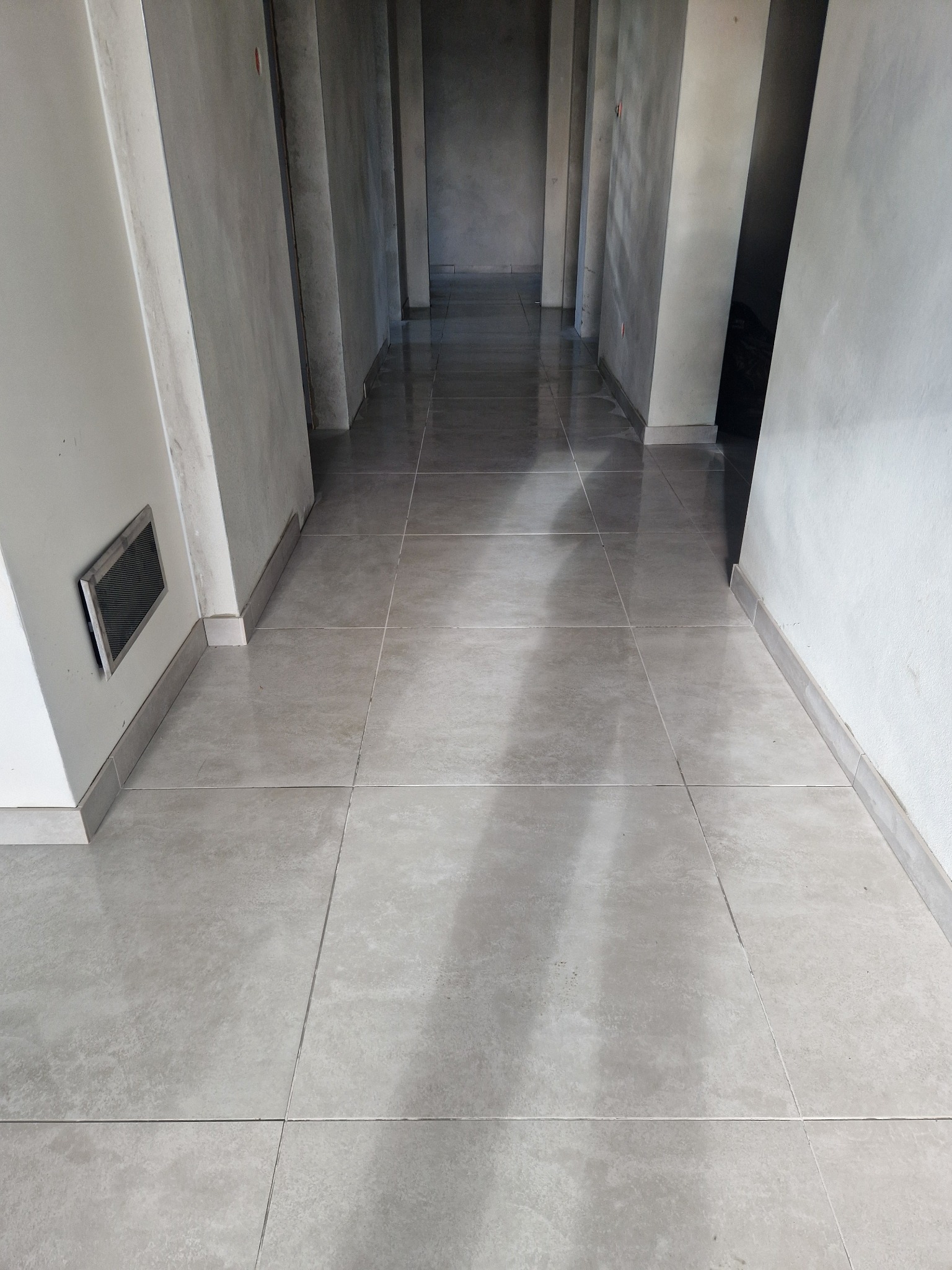 Wyłożona jasnoszarą płytką podłoga w długim korytarzu z surowymi ścianami. Widoczny fragment kratki wentylacyjnej przy podłodze. Perspektywa zbieżna w głąb.