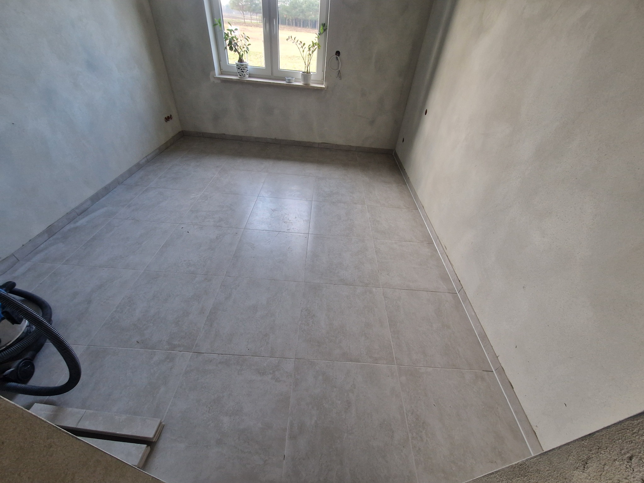 Jasne, kwadratowe płytki podłogowe w pustym pokoju z oknem i roślinami. Ściany w odcieniach szarości, widoczny odkurzacz i zapasowe płytki. Ujęcie z dołu.