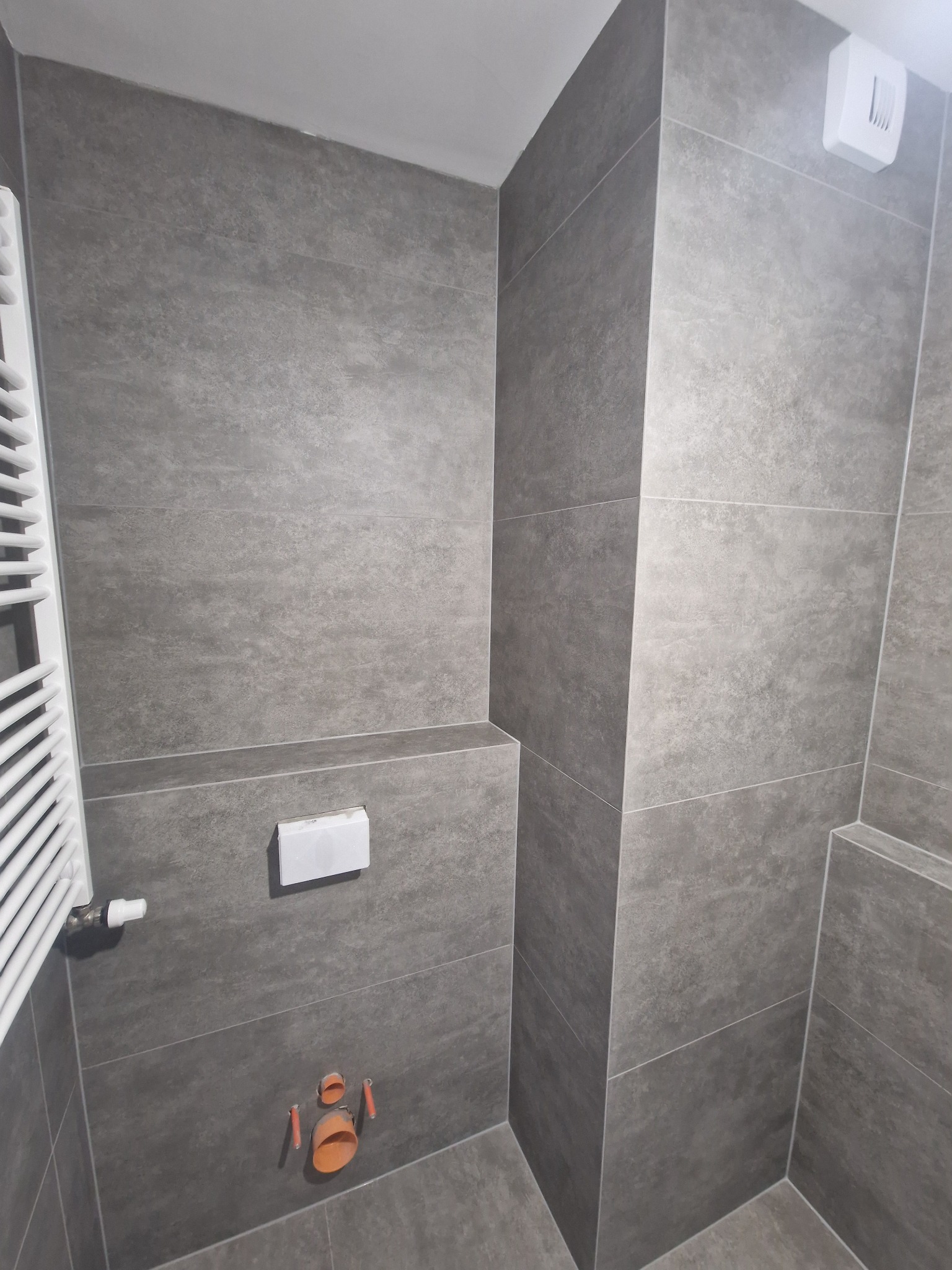 Szara, wykafelkowana łazienka z grzejnikiem drabinkowym, podtynkową spłuczką i otworami na rury. Minimalistyczny design z matowymi płytkami.