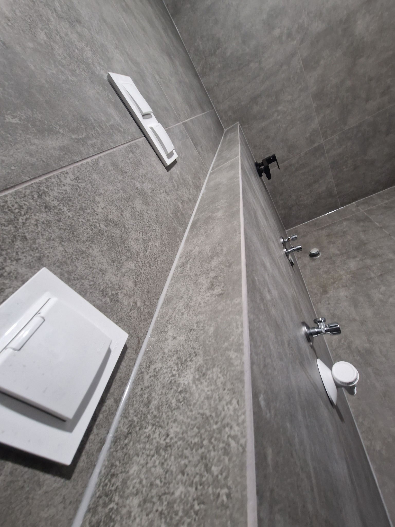 Szara, wykafelkowana łazienka z białymi elementami armatury i włącznikami światła, sfotografowana pod kątem, ukazująca nowoczesny design i dbałość o detale.