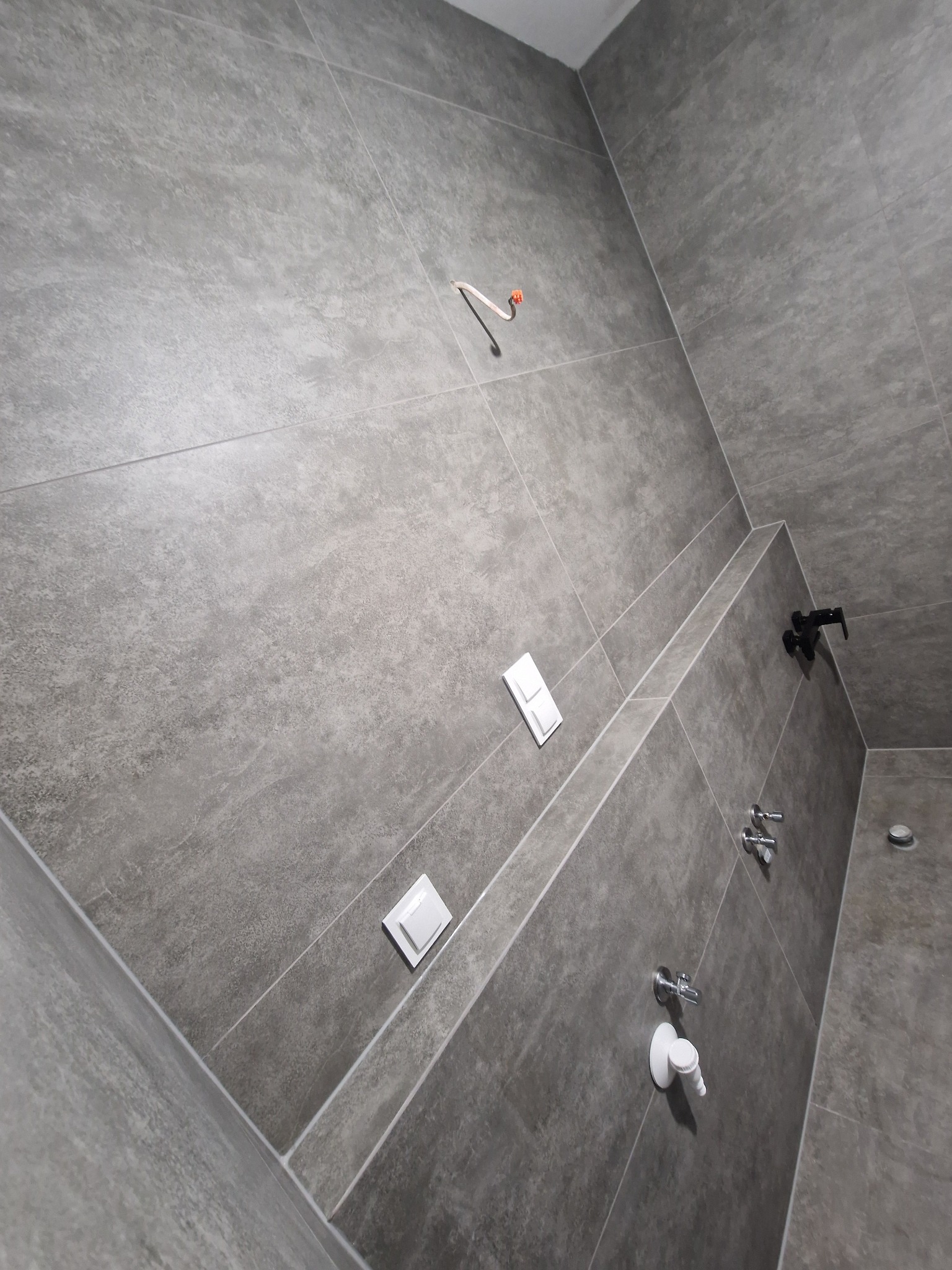 Wykończona łazienka z szarymi, matowymi płytkami na ścianach i podłodze. Widoczne białe włączniki, czarna bateria prysznicowa i chromowane krany. Ujęcie z dołu.