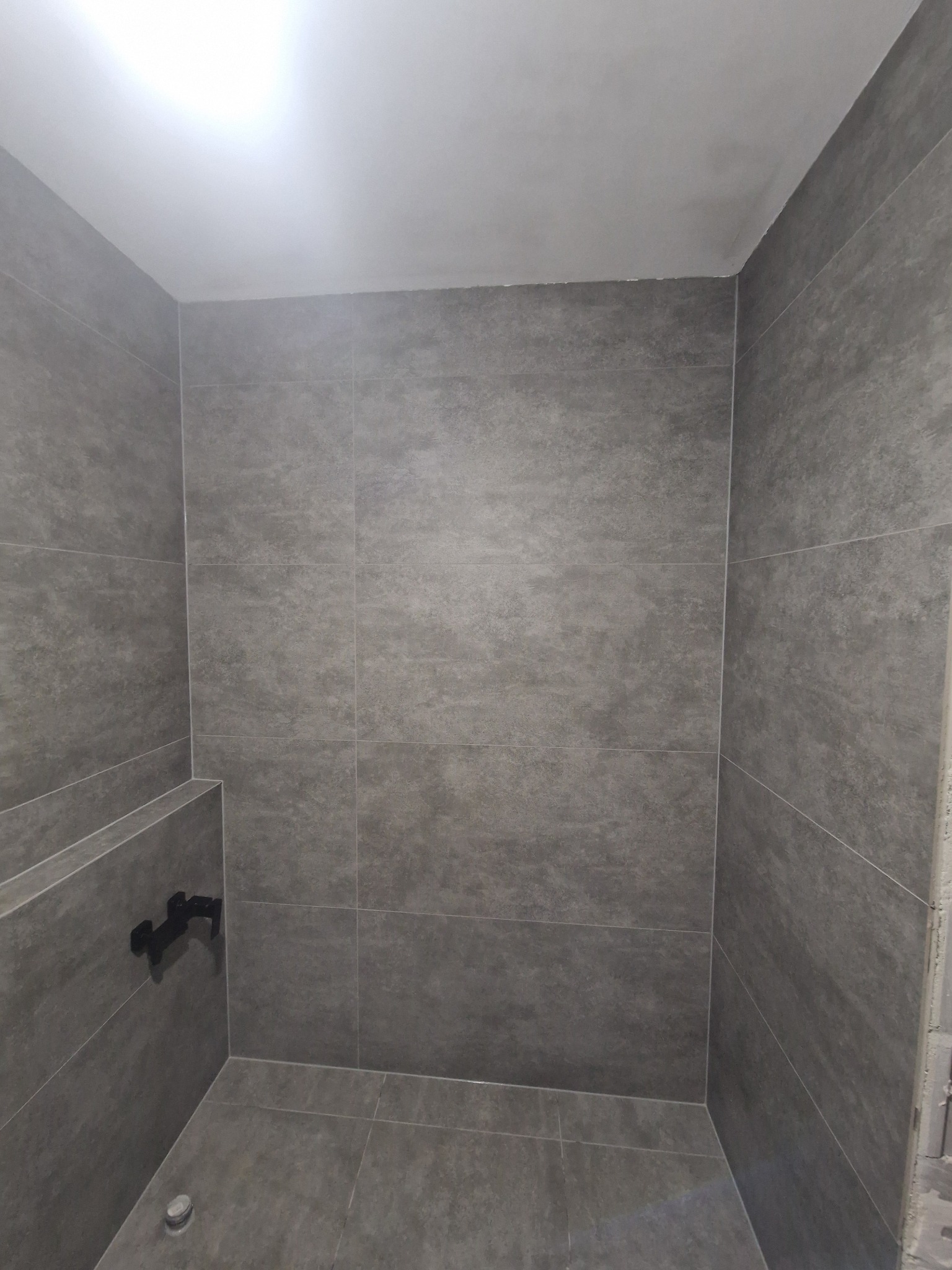 Wykończona kabina prysznicowa z szarymi, matowymi płytkami o nieregularnej fakturze. Widoczna czarna bateria i odpływ w podłodze. Minimalistyczny design.