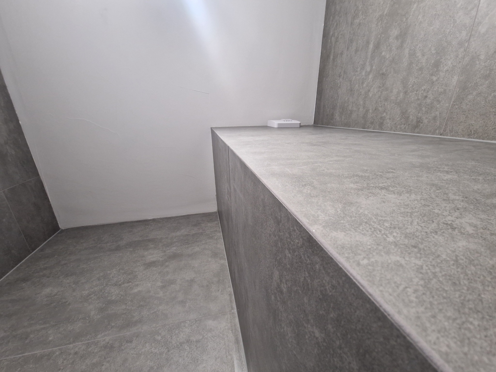 Minimalistyczna łazienka z szarymi płytkami imitującymi beton, z widoczną półką i podłogą. Biała ściana w tle. Nowoczesne wykończenie.