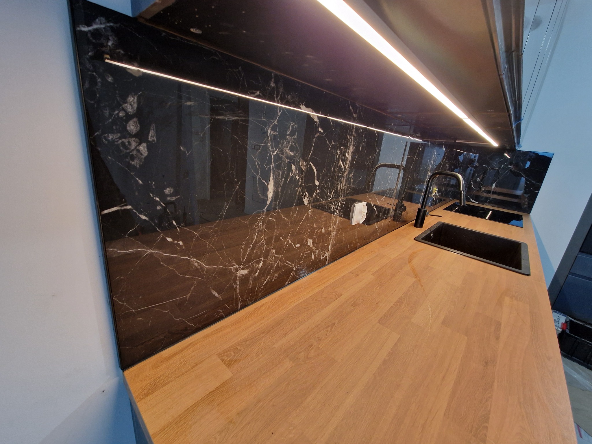 Nowoczesna kuchnia z blatem drewnopodobnym i czarnym panelem ściennym imitującym marmur, oświetlonym listwą LED. Minimalistyczny design.