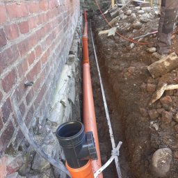 Maciej Sołtys Firma Under Load - Pomarańczowe rury kanalizacyjne w wykopie przy ceglanej ścianie budynku. Widoczne złącza i narzędzia pomiarowe. Częściowo widoczna osoba w roboczym ubraniu.