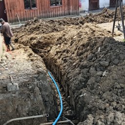 Maciej Sołtys Firma Under Load - Wykop z niebieską rurą wodną w ziemi. Widoczne narzędzia: szpadel, kilof. W tle budynek z drewnianą elewacją. Prace ziemne w toku, instalacja wodna.