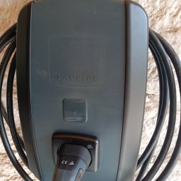 Maciej Sołtys Firma Under Load UnderLoad.biz - Szara ładowarka do samochodu elektrycznego marki Pampere Charge, zamontowana na chropowatej ścianie. Kabel zwinięty wokół urządzenia.