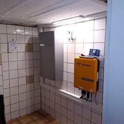 Maciej Sołtys Firma Under Load UnderLoad.biz - Montaż inwertera Solarmax i Enphase na ścianie z kafelkami. Widoczne kable i narzędzia. Notatka z numerami na ścianie. Elementy instalacji fotowoltaicznej w pomieszczeniu.