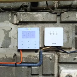 Panel sterowania ogrzewaniem NOXA i AURATON na ceglanej ścianie. Widoczne przewody i gniazdko elektryczne. Ustawienia temperatury i data na wyświetlaczu.