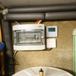 Maciej Sołtys Firma Under Load - Biała skrzynka elektryczna Noark z szarą pokrywą, regulator temperatury Nova i element instalacji grzewczej na tle beżowej ściany i zaizolowanych rur.