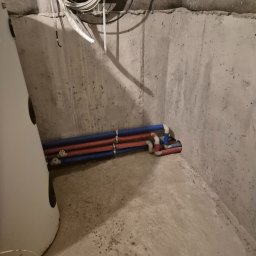 Maciej Sołtys Firma Under Load - Instalacja rur wodnych (czerwone i niebieskie) w betonowej piwnicy, obok białego zbiornika. Widoczne kable elektryczne pod sufitem. Surowy stan wykończenia.