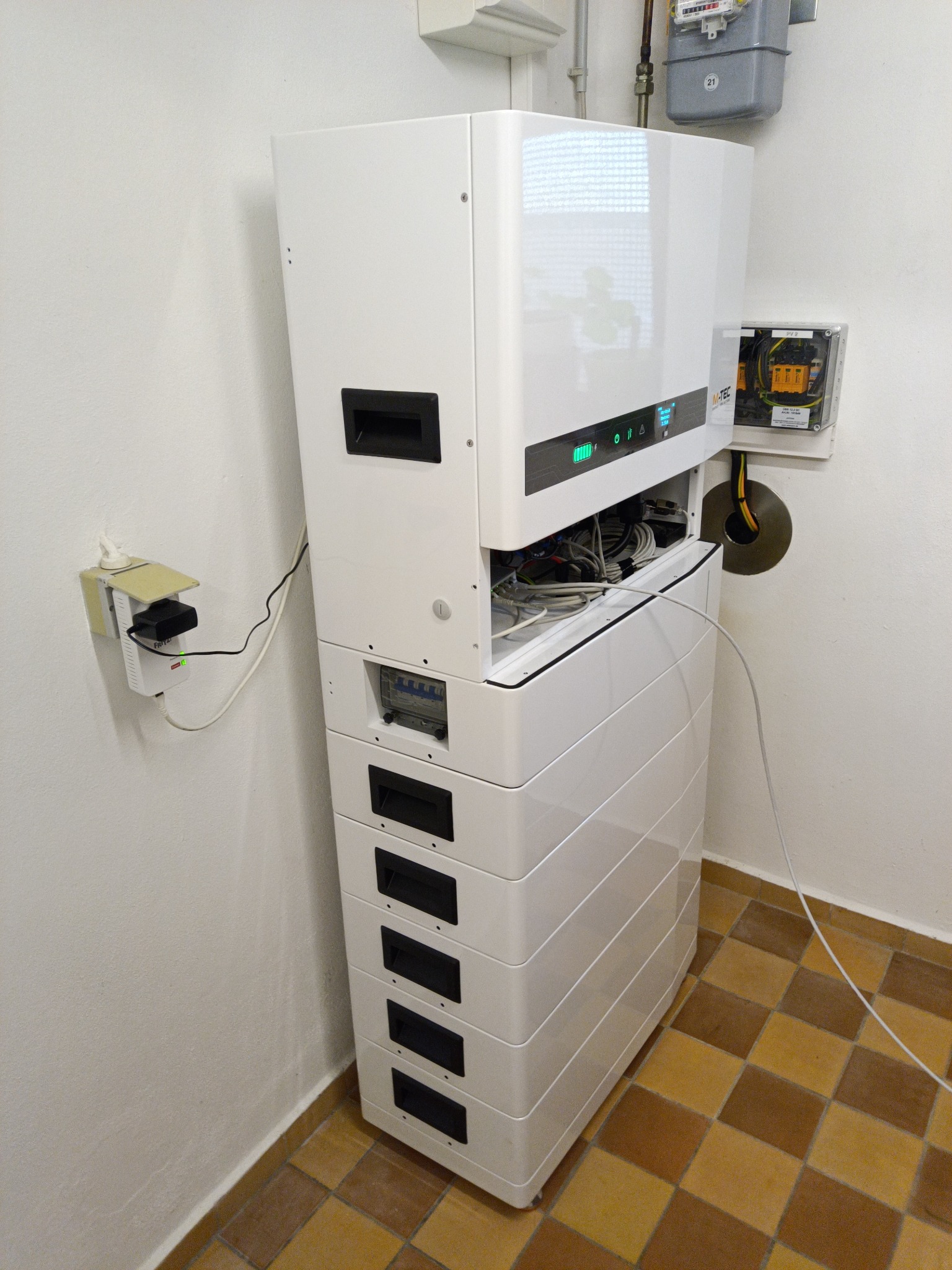 Biały modułowy system zasilania awaryjnego z otwartym panelem, widoczne kable. Na ścianie licznik gazu, gniazdko elektryczne i zabezpieczenia. Brązowa podłoga.