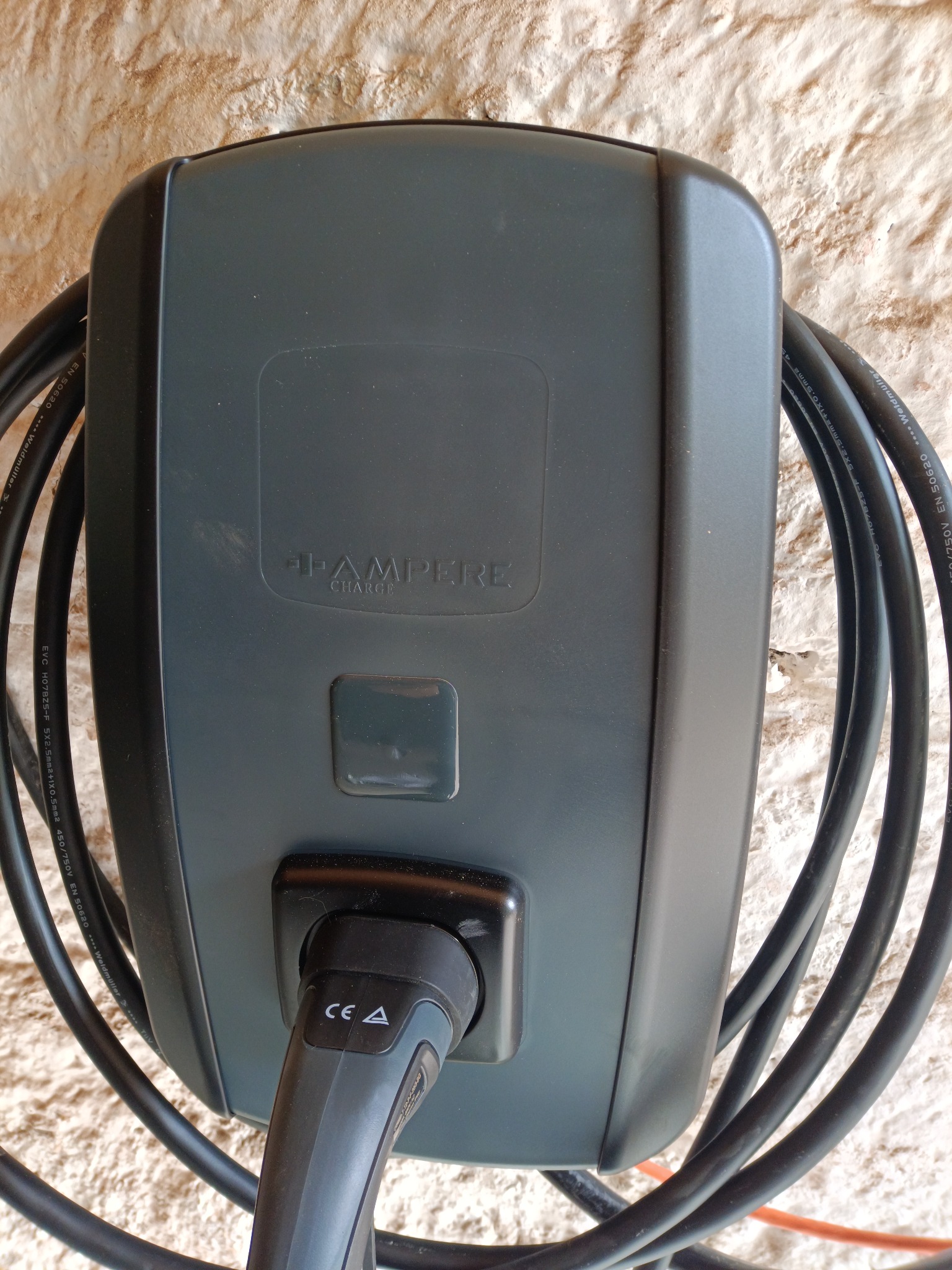 Szara ładowarka do samochodu elektrycznego marki Pampere Charge, zamontowana na chropowatej ścianie. Kabel zwinięty wokół urządzenia.