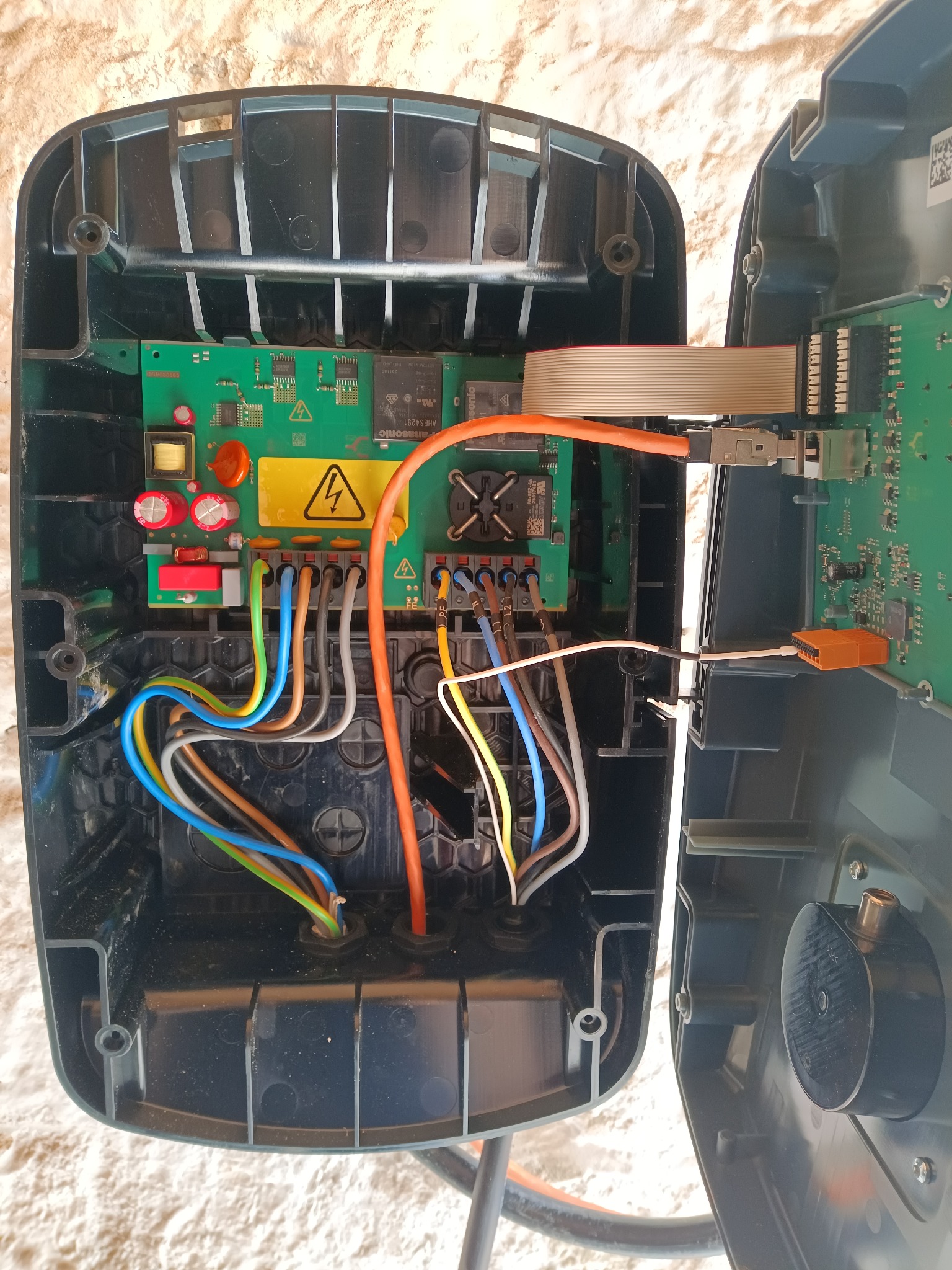 Rozwarte wnętrze stacji ładowania EV z widoczną elektroniką: płytka drukowana, okablowanie w kolorach, złącza. Pomarańczowy kabel zasilający, żółta naklejka ostrzegawcza.