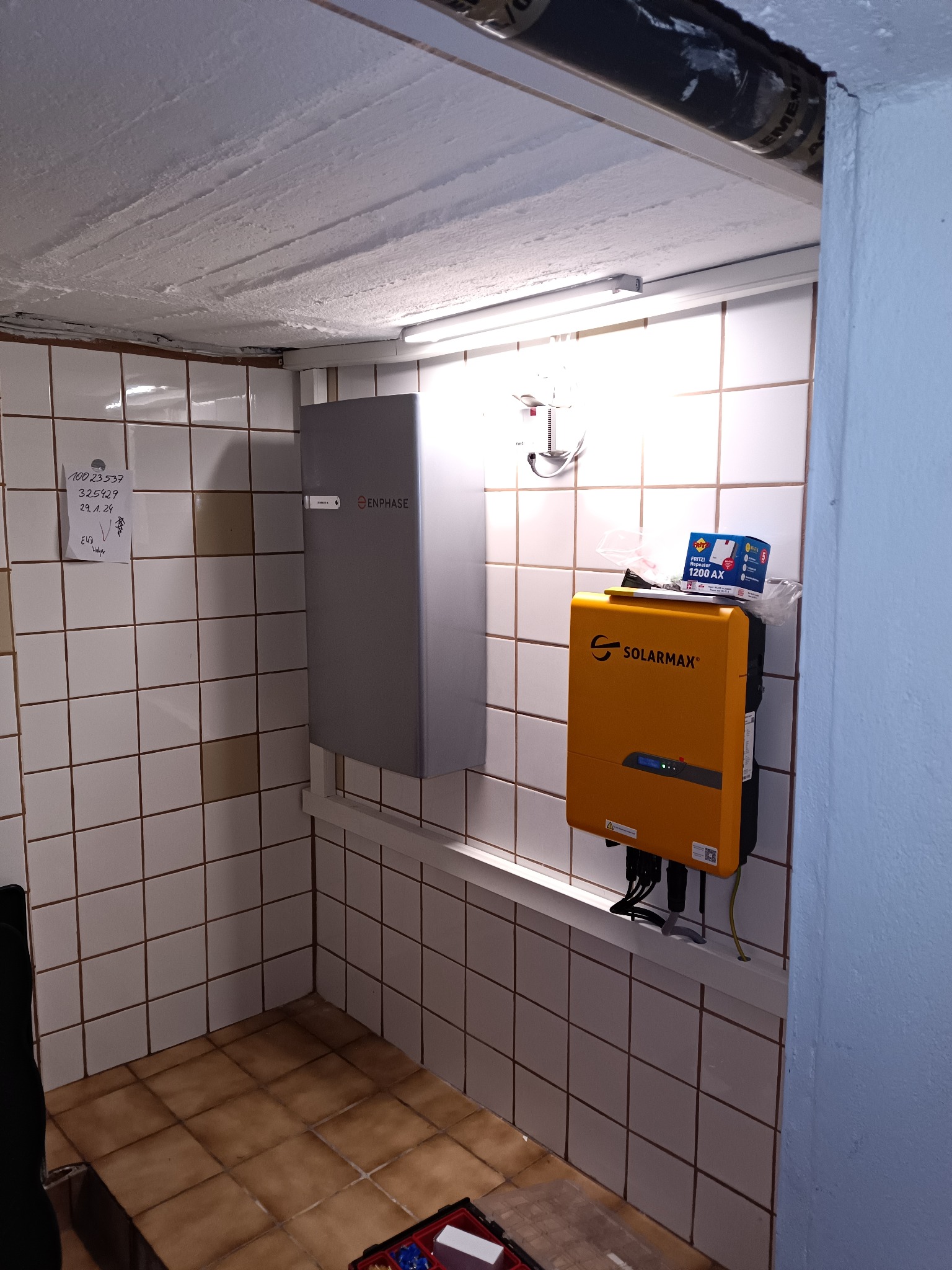 Montaż inwertera Solarmax i Enphase na ścianie z kafelkami. Widoczne kable i narzędzia. Notatka z numerami na ścianie. Elementy instalacji fotowoltaicznej w pomieszczeniu.