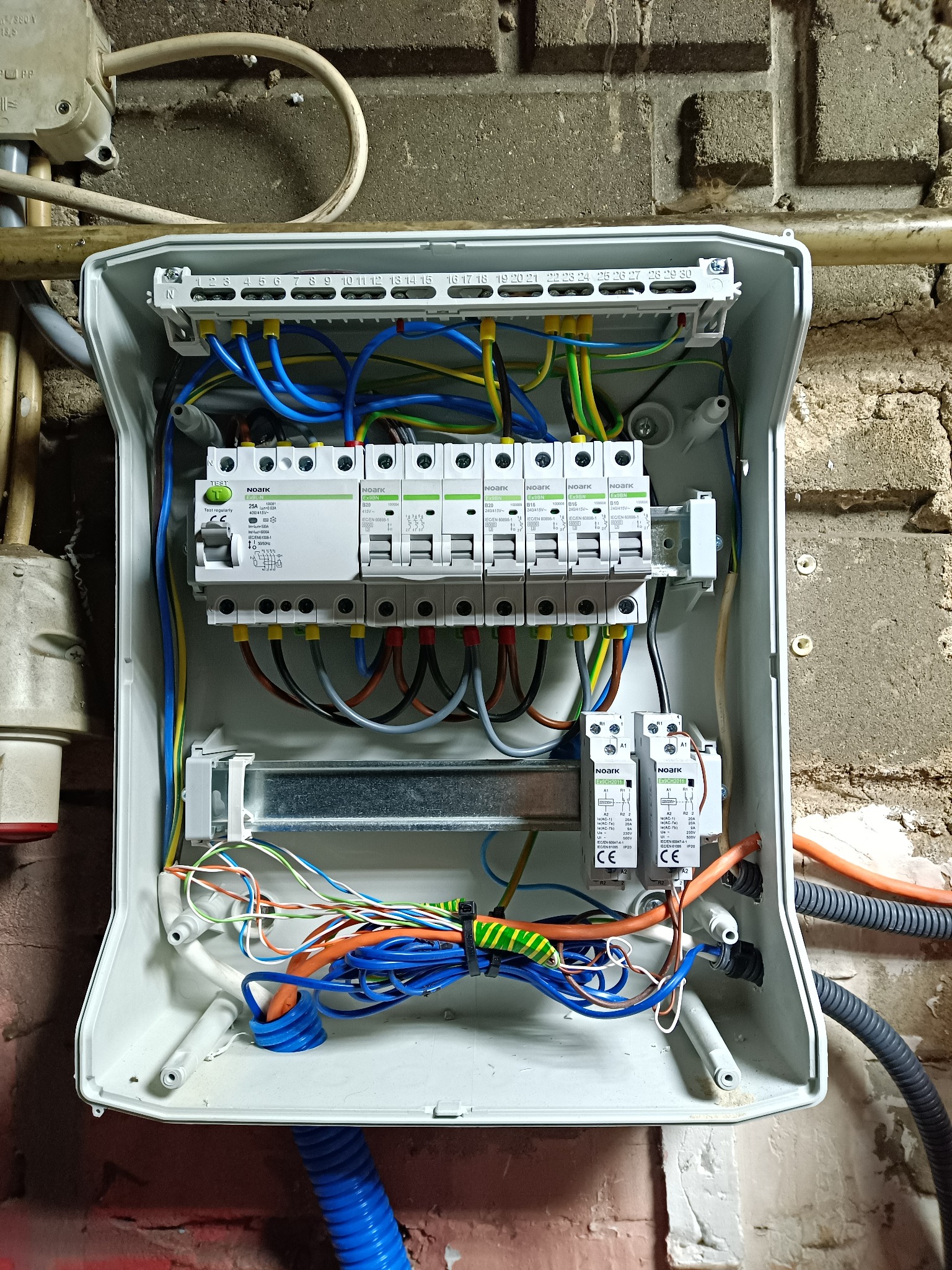 Otwarta skrzynka rozdzielcza z bezpiecznikami i okablowaniem na tle ceglanej ściany. Widoczne przewody w różnych kolorach, listwa zaciskowa i aparatura modułowa.