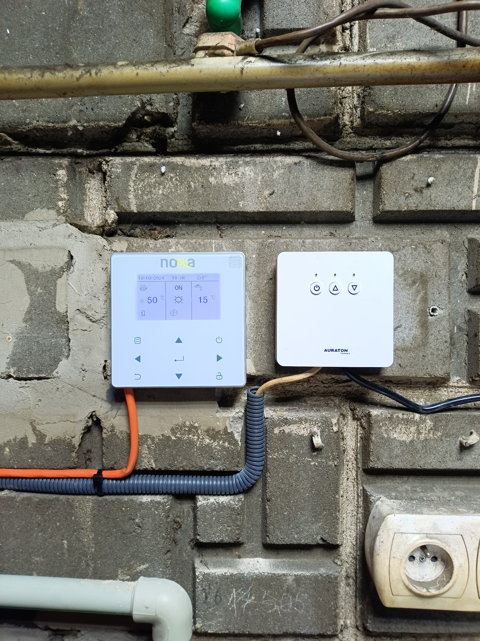 Panel sterowania ogrzewaniem NOXA i AURATON na ceglanej ścianie. Widoczne przewody i gniazdko elektryczne. Ustawienia temperatury i data na wyświetlaczu.