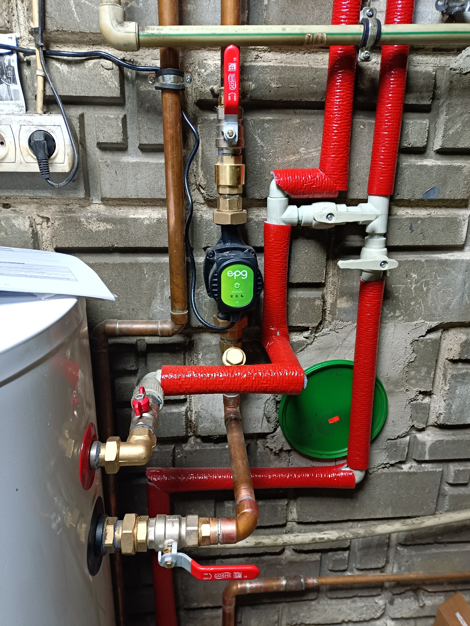 Instalacja grzewcza z rurami miedzianymi i plastikowymi, izolowanymi czerwonym materiałem. Widoczny bojler, pompa obiegowa i zawory na tle szarej ściany z cegły.