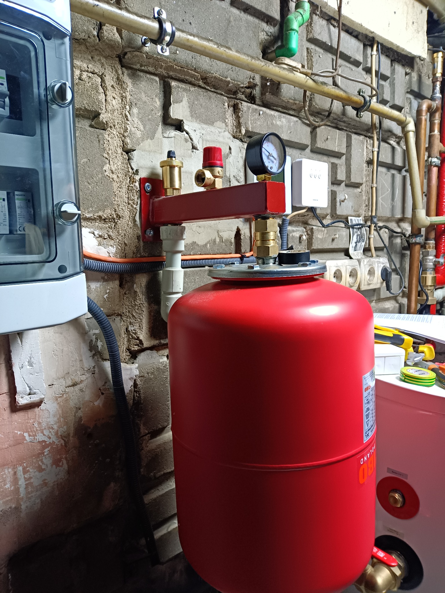 Czerwony zbiornik wyrównawczy CO z manometrem i zaworami, zamontowany na tle ceglanej ściany. Widoczne rury, kable i skrzynka elektryczna.