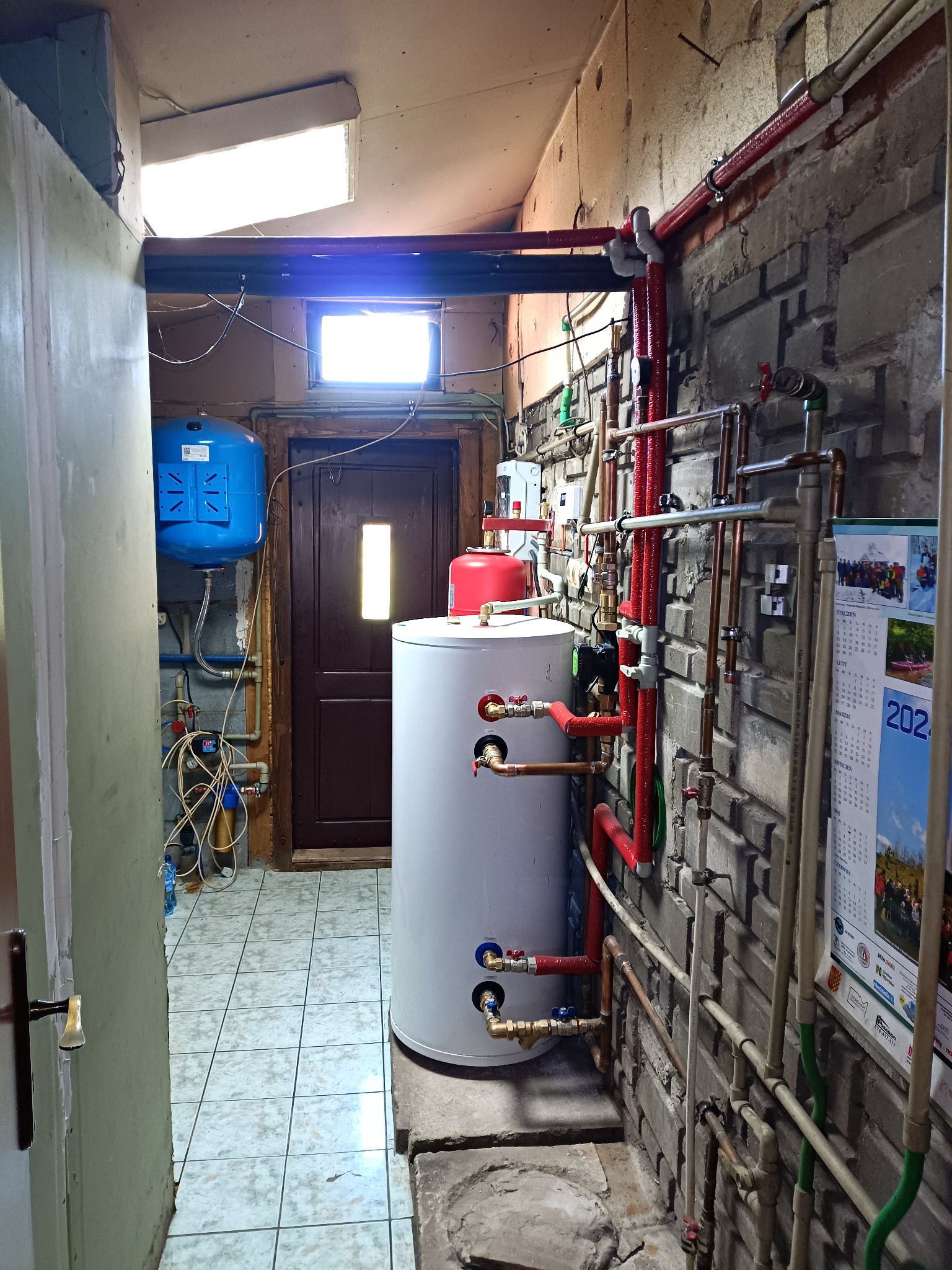 Kotłownia z białym bojlerem, czerwonymi rurami i niebieskim zbiornikiem wyrównawczym na tle surowej, ceglanej ściany. Widoczne liczne instalacje i kalendarz na ścianie.