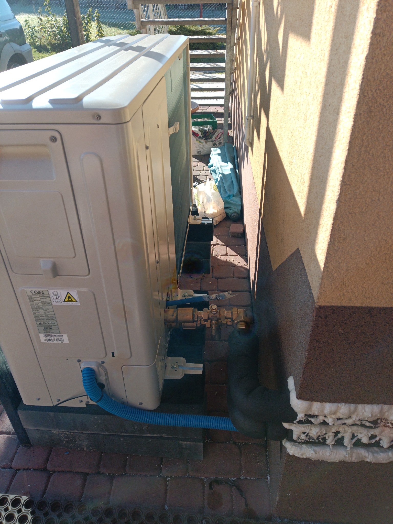 Klimatyzator zewnętrzny zamontowany przy ścianie budynku, widoczne rury i izolacja. Instalacja na ceglanym podłożu, w tle schody i ogrodzenie.
