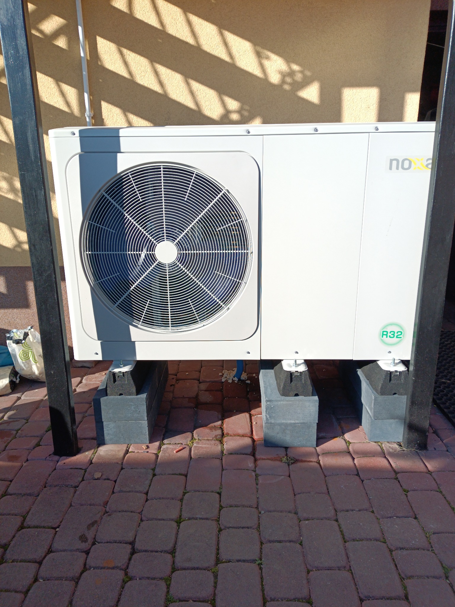 Nowoczesna, biała pompa ciepła Noxa R32 zamontowana na betonowych bloczkach na tle ceglanej kostki brukowej i ściany z cieniem. Widoczne elementy instalacji i konstrukcja wsporcza.