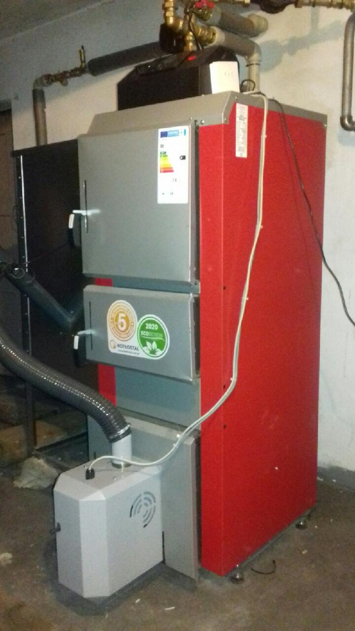 Czerwono-szary piec na paliwo stałe marki Kotłostal, z widocznymi naklejkami '5' i '2020 Ecodesign', podłączony do instalacji w kotłowni.