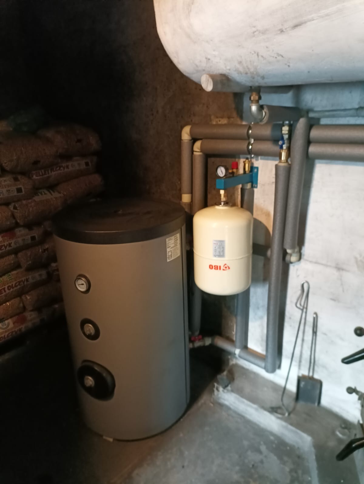 Szary bojler z białym zbiornikiem wyrównawczym IBO, rury z izolacją termiczną, worki z pelletem w tle. Elementy instalacji grzewczej w kotłowni.