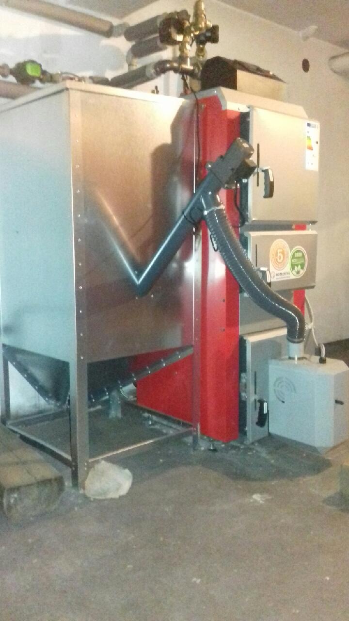 Nowoczesny piec na paliwo stałe z podajnikiem, z widocznymi elementami instalacji. Czerwono-srebrna kolorystyka, szare rury i betonowa podłoga w tle.