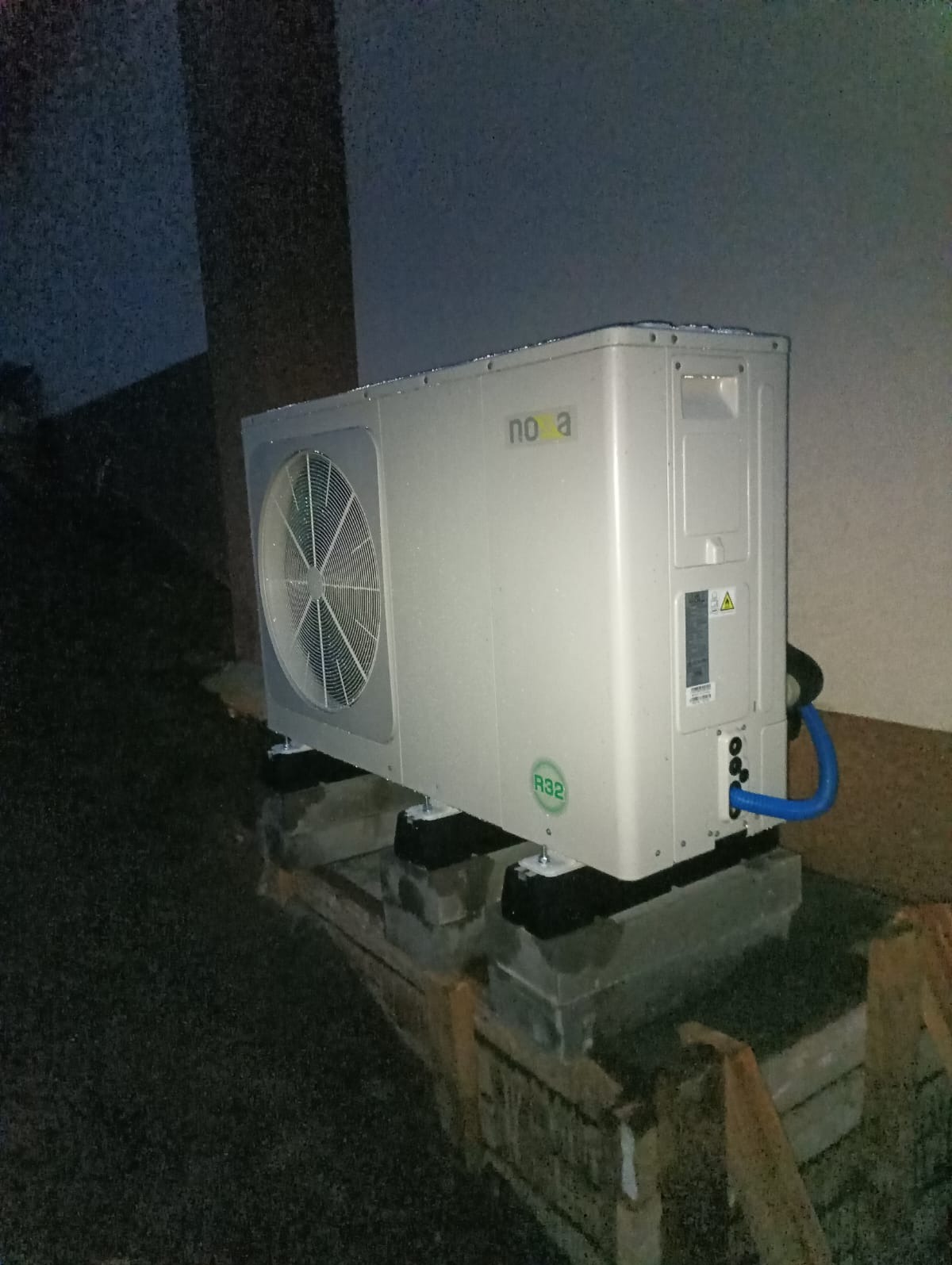 Biała pompa ciepła NOVA R32 zamontowana na betonowej podstawie z widocznym niebieskim przewodem, na zewnątrz budynku, nocą.