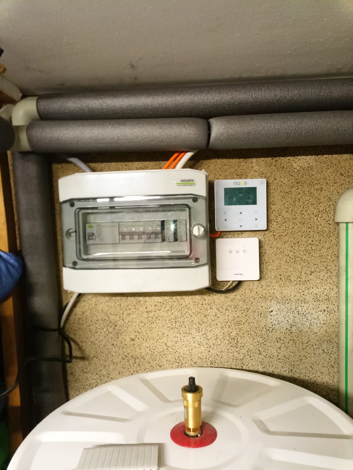 Biała skrzynka elektryczna Noark z szarą pokrywą, regulator temperatury Nova i element instalacji grzewczej na tle beżowej ściany i zaizolowanych rur.