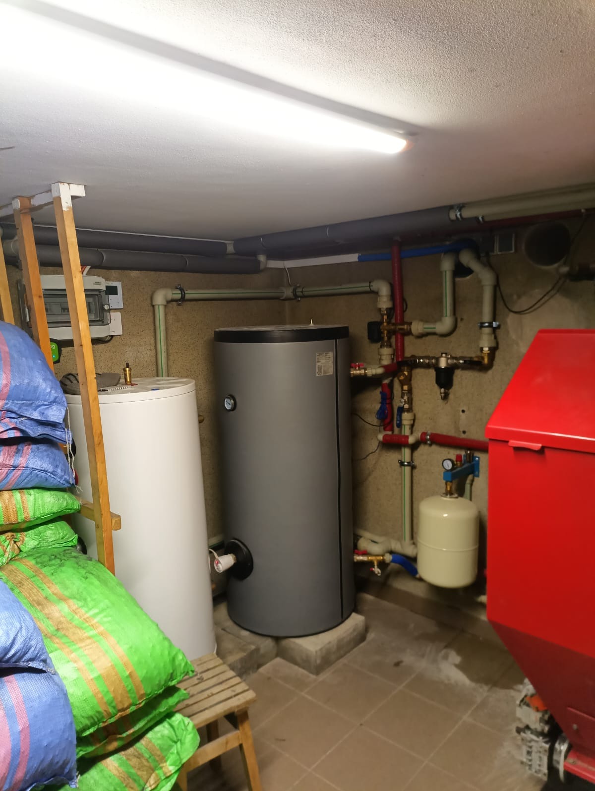 Instalacja CO z piecem na paliwo stałe w piwnicy w Łapszance. Widoczne zbiorniki, rury i zawory. Na pierwszym planie worki z ekogroszkiem.