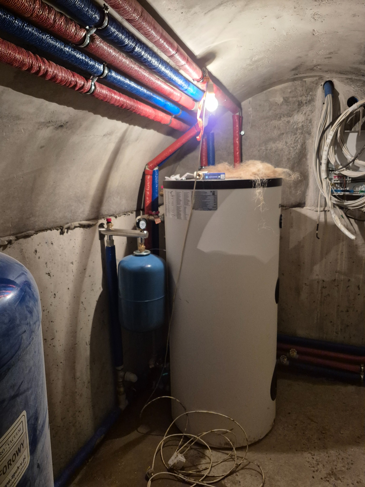Bojler z podłączonymi rurami w izolacji termicznej, manometr, zbiornik wyrównawczy i przewody elektryczne w piwnicy ze sklepieniem łukowym.