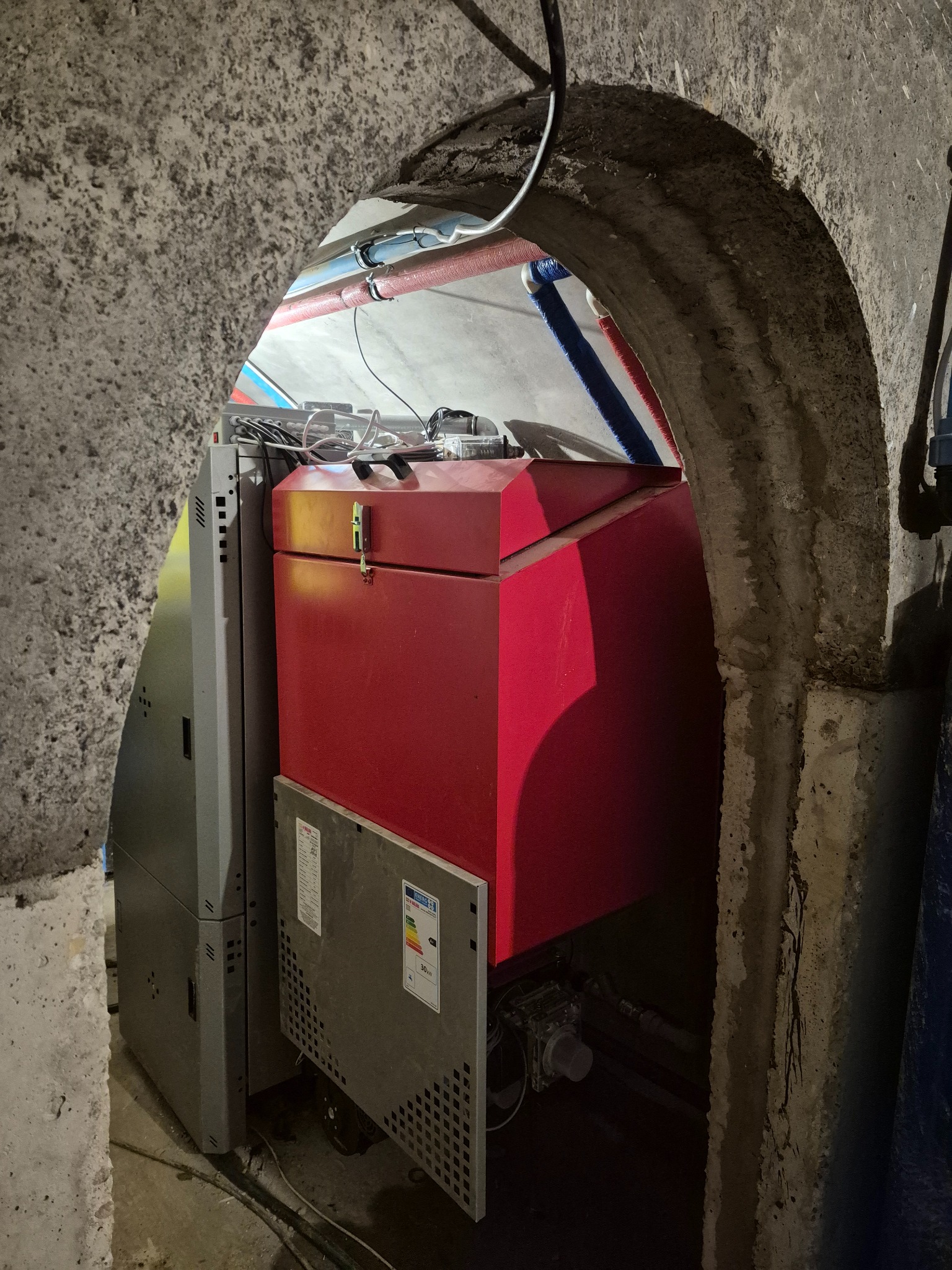 Czerwony piec na paliwo stałe w betonowej wnęce z widocznymi rurami instalacji. Szary panel boczny pieca, etykieta energetyczna. Surowe wykończenie wnęki.